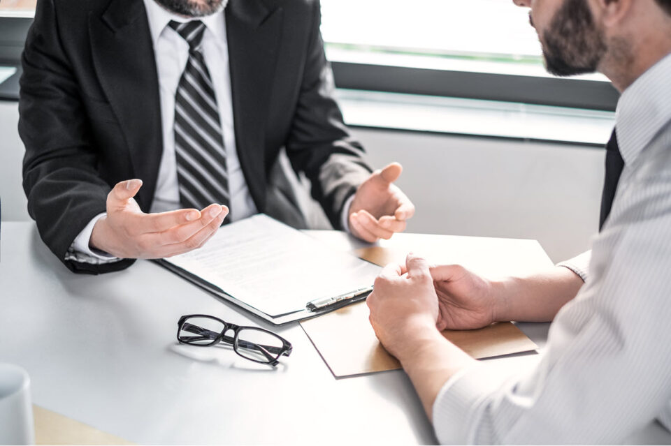Two businessmen in formal attire having a discussion at a desk with documents, a clipboard, and eyeglasses