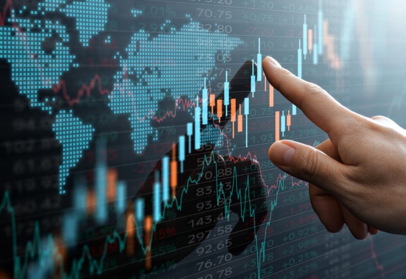 Hand pointing at a digital screen displaying global financial data, stock market charts, and a world map overlay